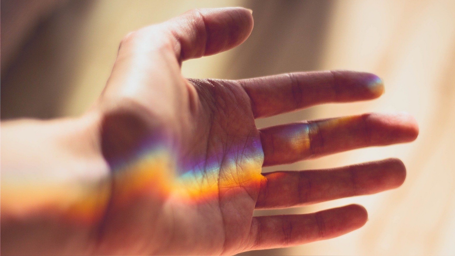 Eine Hand ist offen im Licht, ein Regenbogenschimmer gleitet über die Haut. Die Geste wirkt einladend, fragil und voller stiller Bedeutung – wie ein Ja zum Berührtwerden.