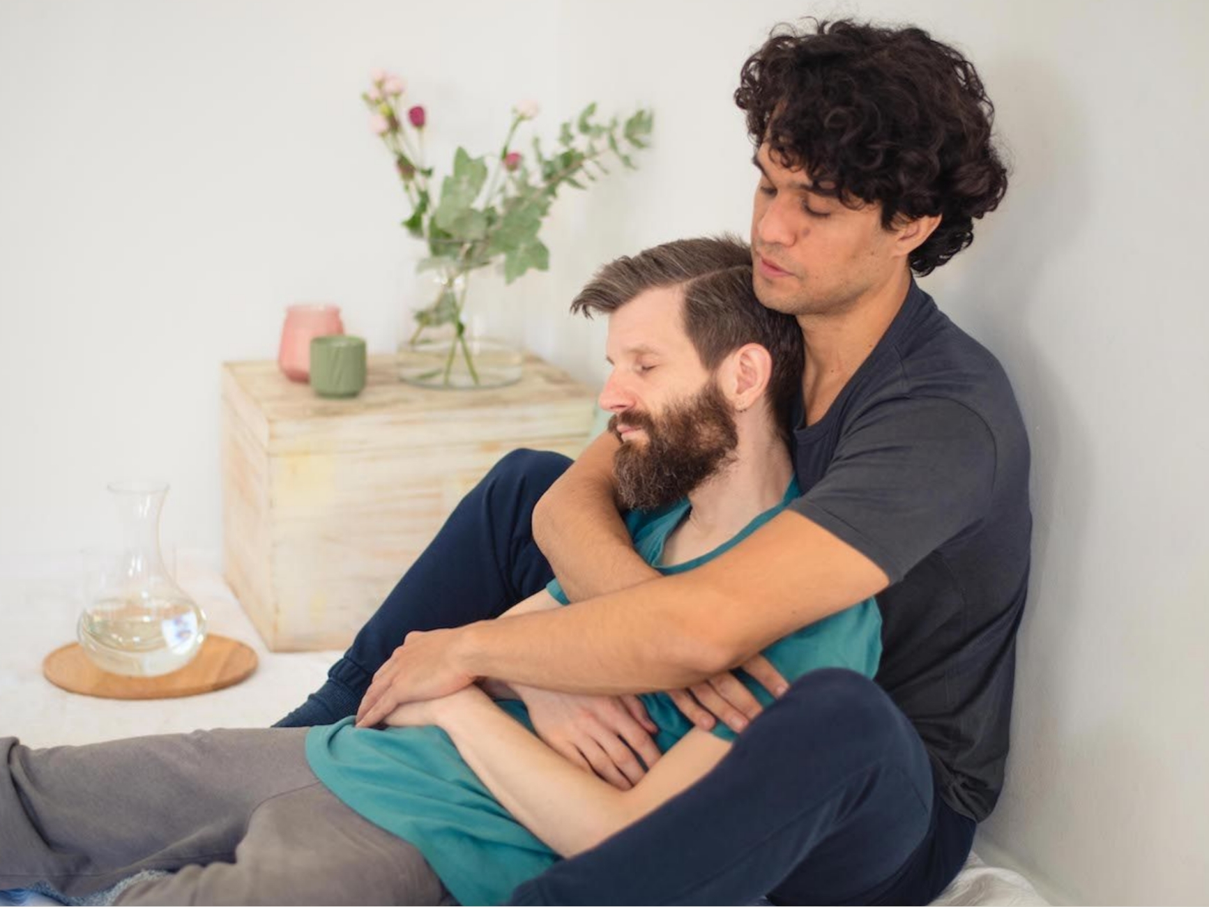 Eine Person sitzt hinter einer anderen und hält sie in einer aufrechten, ruhigen Umarmung.